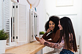 Side view of content diverse female friends sitting at counter in bar and using smartphones while enjoying weekend together