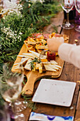 Anonymous guest reaches for a selection from a bountiful cheese platter amidst a floral banquet, with glasses of wine and cheerful ambiance