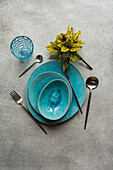 Top view of a vibrant table setting featuring turquoise ceramic dishes and cutlery paired with a decorative glass and potted plant on a textured surface.