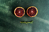 Two halves of juicy blood oranges with a sprig of rosemary on a textured green background