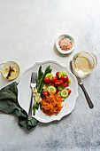 Top view of a well arranged vegetable plate with asparagus spears, sliced tomatoes and cucumbers, grated carrot, cheese, beside a glass of wine and a dish of pink salt.