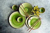 Top view of a bright dinner setting featuring green ceramic plates, bowls, and cups with wooden chopsticks on a textured surface.