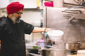 A chef in a red turban carefully adds ingredients to a steaming pot in a professional kitchen