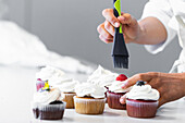 Unrecognizable crop baker with silicone brush spreading sweet syrup on berries while preparing vegan cupcakes in kitchen