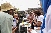 A lively group of friends engages in conversation while enjoying a BBQ and music outdoors. Several are looking at each other, and some look away, creating a relaxed and social atmosphere.