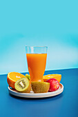 Eine Vielzahl frischer Früchte, darunter Orangen, Kiwi und ein Glas Orangensaft, kunstvoll auf einem Teller vor einem auffallend blauen Hintergrund angeordnet.