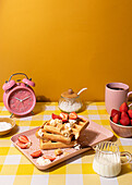 Ein gemütliches Morgenarrangement mit einem Teller belgischer Waffeln mit Sahne und Erdbeeren sowie einem rosa Wecker, einer Kaffeetasse, einer Zuckerdose und einem Milchkännchen auf einer gelb karierten Tischdecke