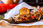 Gourmet dish featuring a stuffed eggplant with cherry tomatoes, bell peppers, onions, garlic, and potato slices, topped with cheese, served with a garnish of microgreens.