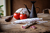 Vintage kitchen setting featuring a sliced cured sausage, whole salami, ripe tomatoes, and rustic pottery.