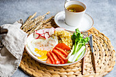 A nutritious lunch setup featuring fresh organic vegetables including reddish, tomato, and celery sticks, accompanied by a fried egg and bulgur cereal, served with a cup of fragrant tea.