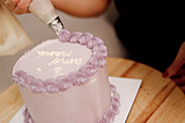 A baker applies intricate purple icing to a cake using a piping bag, set on a wooden table, showcasing the art of cake decorating