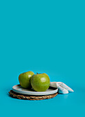 Two vibrant green apples neatly placed on a gray plate over a woven round mat. This composition showcases a simplistic yet appealing approach to presenting healthy and wholesome food, emphasized by a striking blue background.