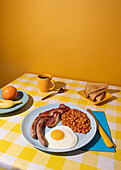 Yellow coloured Tablecloth and background with a delicious full English Breakfast, egg, beans, sausages and bacon, some toasted bread slices, fruit and a cup of coffee