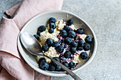 A nourishing breakfast featuring a bowl of cottage cheese topped with organic blueberry jam and fresh blueberries, accompanied by a spoon on a light gray surface with a pink napkin