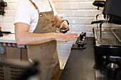 A barista in an apron holds a portafilter filled with ground coffee, ready to brew with an espresso machine in a cafe setting.