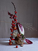 An artful arrangement featuring tiny red crab apples on branches, supported by a swede, mushrooms, radicchio leaves, and delicate white flowers, set against a soft gray backdrop.