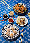 From above, a vibrant display of various traditional Chinese dishes served on a blue patterned tablecloth. Includes dim sum, noodles with shrimp, and accompanying sauces