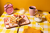 Eine reizvolle Frühstücksszene mit einem Tablett knuspriger Waffeln, belegt mit frischen Erdbeeren und Schlagsahne, ergänzt durch eine Tasse Kaffee, ein Kännchen Milch und einen vibrierenden rosa Wecker auf einem leuchtend gelben Hintergrund