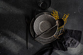 Top view of a sophisticated black table setting featuring dark plates, utensils, and a yellow mimosa garnish.