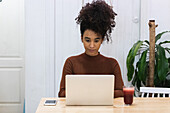 Focused African American female freelancer sitting at table with refreshing smoothie and browsing netbook while working on project in cafe