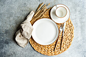 From above, a neat table setting features bright ceramic dinner plates, a cup, shiny cutlery, a woven placemat, and wheat sprigs on a textured surface.