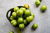 A top view of fresh, organic green plums arranged in a dark ceramic bowl set on a textured grey surface, hinting at Georgian, Turkish, and Greek culinary traditions