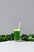 A vibrant glass of green kale juice paired with fresh curly kale leaves on a neutral background, featuring a yellow-striped straw for a fresh aesthetic