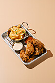 A tray containing golden fried chicken wings paired with seasoned fries, complete with creamy and tangy dipping sauces presented on a beige background.