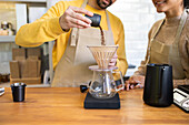 A barista in a yellow sweater pours ground coffee into a pour-over maker while colleague watches with interest, in a cozy cafe setting.