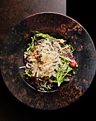 A delicious gourmet salad topped with shaved cheese, served on a textured, rustic-style plate against a dark background, perfect for a healthy meal