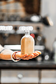 A cropped unrecognizable person's hand holding a clear bottle of homemade liqueur with a soft focus kitchen background, showcasing the art of craft distillation.