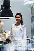 Content female baker in uniform standing behind showcase in bakehouse and looking away