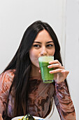 Delighted female sitting in restaurant and drinking refreshing green smoothie while enjoying weekend and looking away