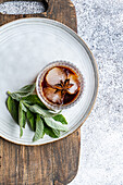 Top view of a glass of cherry cocktail with ice cubes and a sprig of mint, on a rustic serving board with a white plate.