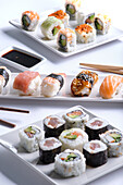 An enticing assortment of sushi rolls neatly arranged on two white plates, accompanied by soy sauce and chopsticks.