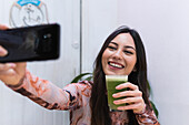 Positive Frau sitzt mit einem Smoothie am Tisch und macht ein Selfie mit ihrem Handy, während sie in einer Bar am Wochenende chillt