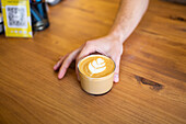 A person's hand gently grips a cup of latte with a foam art design on a wooden surface, portraying a relaxed coffee moment.