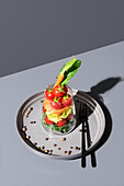 Top view of a vibrant layered vegetable salad served in a transparent glass placed on a metal tray with a gray background, accompanied by a black fork.