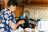 Eine ältere Frau wird beim Abwaschen von Geschirr in ihrer heimischen Küche aufgenommen, umgeben von Kochutensilien und einem gefliesten Vintage-Hintergrund