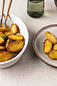 A top view image showcasing golden roasted potatoes sprinkled with herbs, presented on a stylish white bowl and grey ceramic plate.