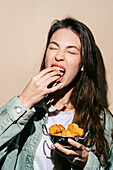 A cheerful young woman in a casual outfit biting into a golden fried mozzarella bite with enjoyment, holding a bowl of additional pieces.