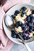 Genießen Sie ein nahrhaftes Frühstück mit saurer Sahne, Bio-Blaubeermarmelade und frischen Blaubeeren auf cremigem Hüttenkäse, serviert in einer geschmackvollen Keramikschale mit einem alten Löffel