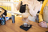 A barista with a black kettle pours hot water over coffee grounds in a glass dripper while a customer waits.