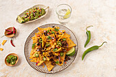 Top view of a plate of nachos generously topped with melted cheese, ground beef, jalapenos, and fresh herbs, paired with guacamole and lime on a light textured surface.