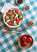 Ein bunter griechischer Salat in einer weißen Schüssel auf einer blau-weiß karierten Tischdecke, komplett mit Tomaten, Gurken, Fetakäse und Oliven, dazu ein Salzstreuer und frische Tomaten auf der Seite