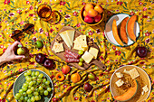 Aerial view of an anonymous hand in a colourful summer picnic setting, with a wooden cheese board laden with various cheeses, crackers and salami, surrounded by fresh fruit such as grapes, apples and melons on a flowery yellow blanket