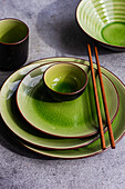 From above view of a stylish table setting featuring bright green ceramic plates and bowls, complemented by wooden chopsticks, all arranged on a textured gray surface.