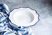 A pristine white plate with a scalloped blue edge rests on a striped cloth, presented on a textured grey surface