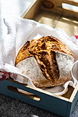 A fresh loaf of artisanal sourdough bread made from rye flour, presented in a wooden tray with a linen cloth, showcasing a rustic kitchen theme