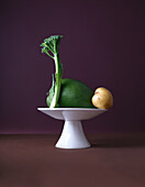 A creative display featuring a mango, potato, and broccoli stalk artfully arranged on a small, elegant white pedestal against a rich purple background